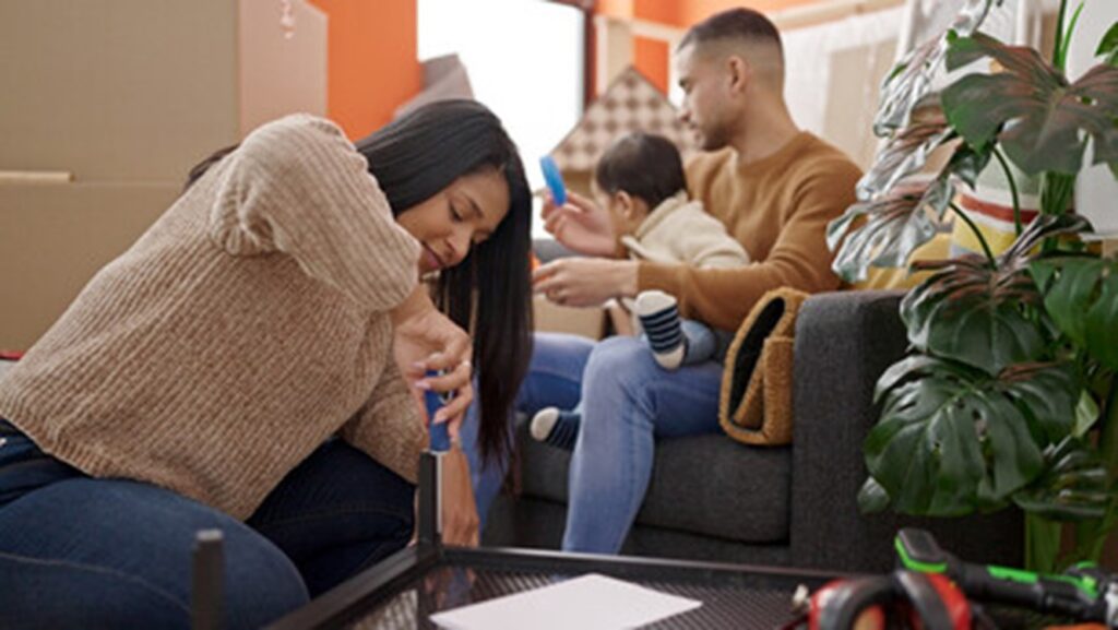 A woman using a screwdriver on the table as man and baby sit in the background